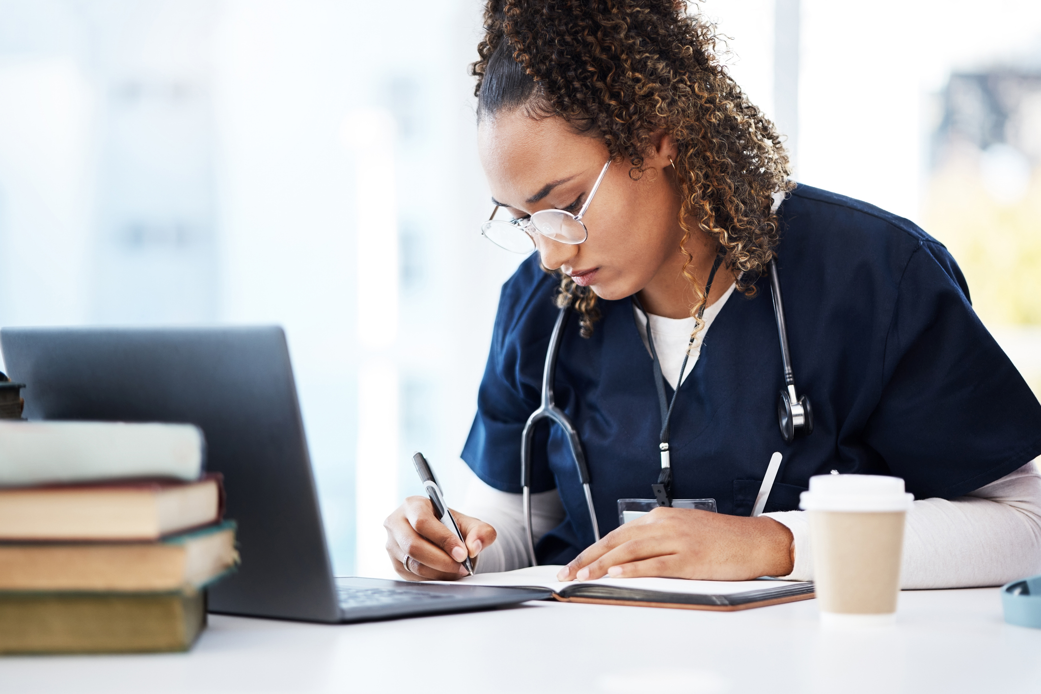 Uma jovem mulher com cabelos cacheados e óculos, vestindo um uniforme médico azul (scrubs) e com um estetoscópio ao redor do pescoço, está sentada em uma mesa. Ela está concentrada, escrevendo em um caderno aberto com uma caneta. Em frente a ela, há um laptop aberto e uma pilha de livros