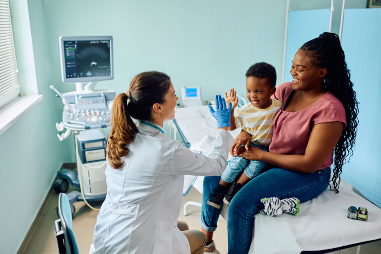 Médica com luvas azuis interagindo com um menino sorridente sentado no colo da mãe em um consultório pediátrico.