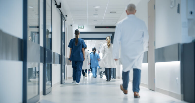 Profissionais de saúde caminham por corredor de hospital moderno, usando jalecos e uniformes clínicos.