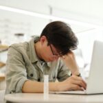 Jovem com expressão de cansaço e sinais de burnout, apoiando a cabeça enquanto trabalha no notebook em ambiente interno.