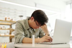 Jovem com expressão de cansaço e sinais de burnout, apoiando a cabeça enquanto trabalha no notebook em ambiente interno.