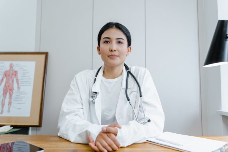 Médica jovem com jaleco e estetoscópio sentada à mesa em consultório, olhando para a câmera com expressão serena e profissional.