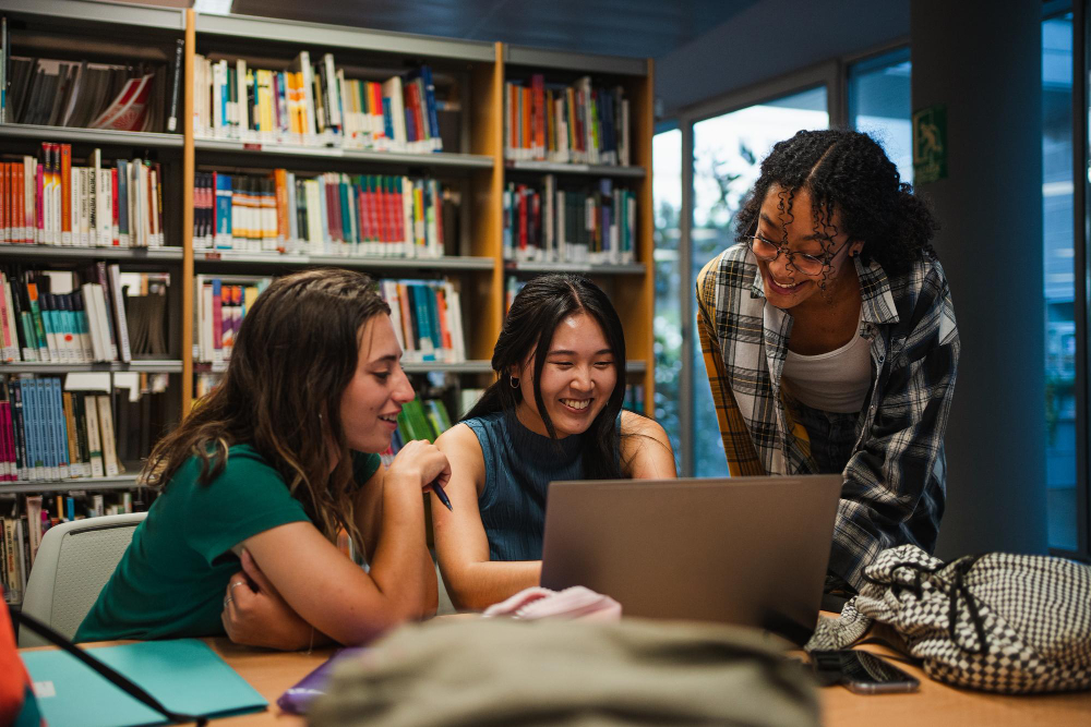 Três estudantes de Medicina estudam juntas em uma biblioteca, reunidas em torno de um notebook durante a semana de provas.