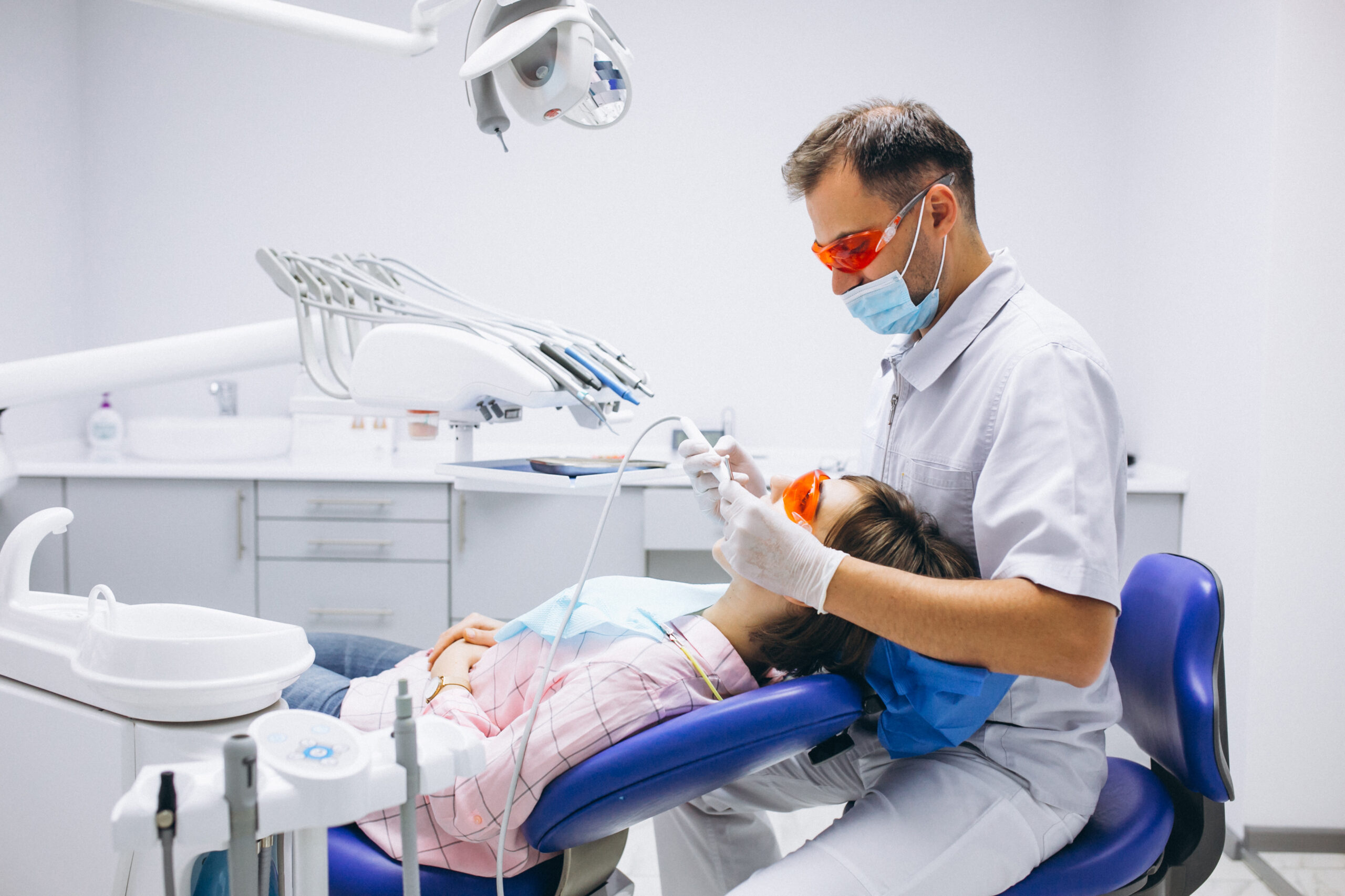 Dentista realizando atendimento em paciente em consultório moderno, ambos usando óculos de proteção e equipamentos de segurança.