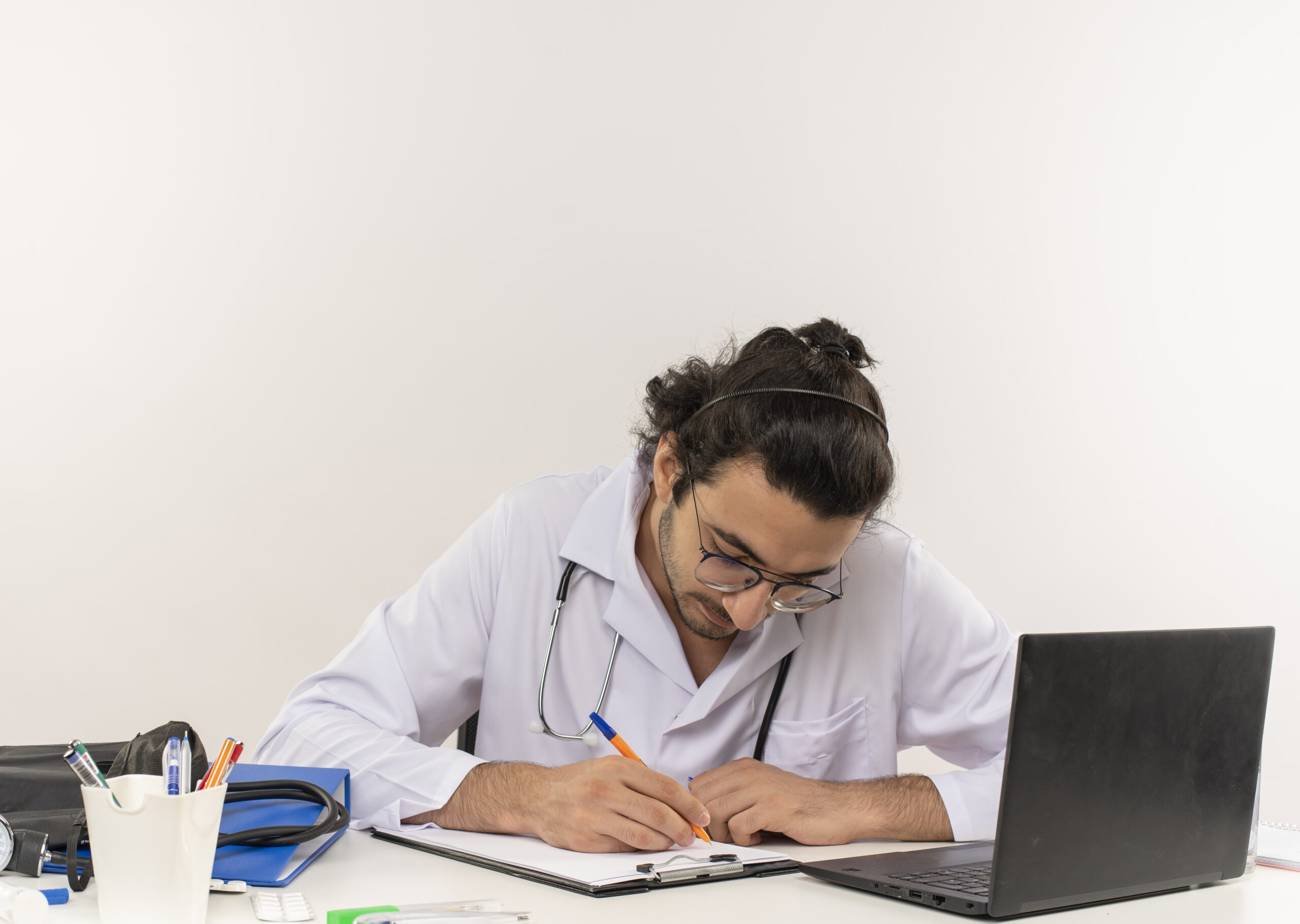 Médico jovem com jaleco e estetoscópio escreve em uma prancheta sobre uma mesa, ao lado de um notebook e itens médicos.